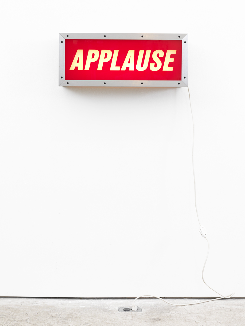 A sculpture of a wall aluminum lightbox with the word "APPLAUSE" in white slanted lettering on a red background and a white cord hanging down from the right side, mounted on a white wall.
