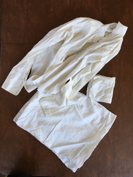 A white karate gi, consisting of a jacket and pants, is laid out on a dark wooden surface. The gi is slightly wrinkled and unfolded, displaying its traditional simple design.
