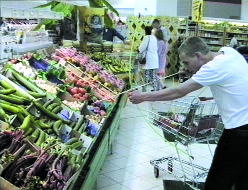 A video still shows the artist, a young white man in a white t-shirt, aiming a bow and arrow at a display of assorted vegetables in a grocery store.