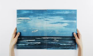 A color photograph of two light-skinned hands holding a book open to an ocean view.