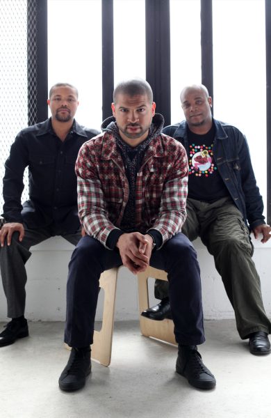 Three men sitting in front of a bright white window look directly at the camera