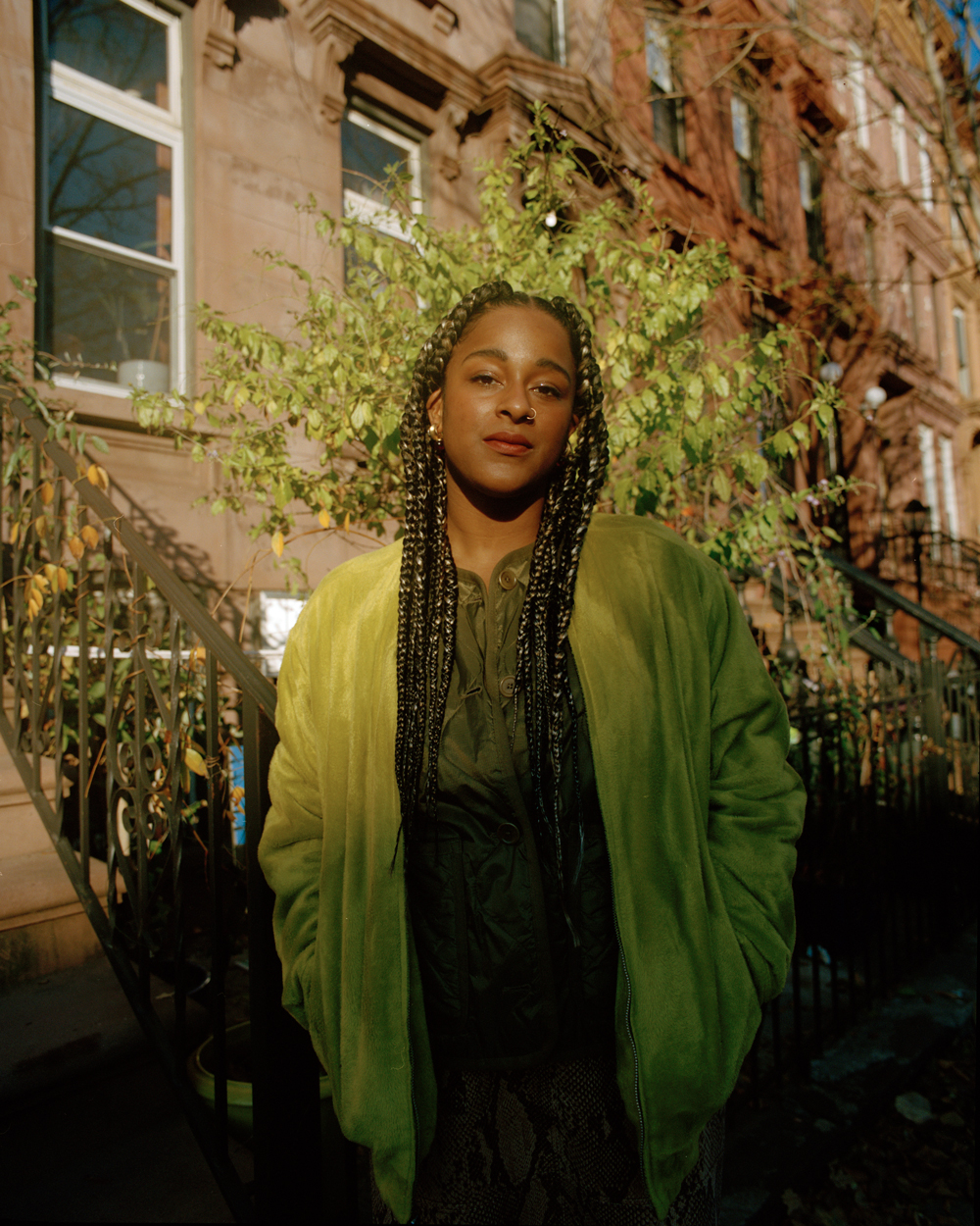 Photo of a person with medium dark skin and long braids standing against brownstone apartments