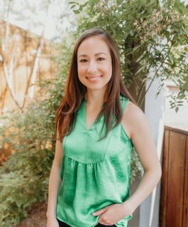 A person stands outdoors, smiling, wearing a sleeveless green top and dark pants. Behind them are green leafy plants and a wooden fence. The lighting is natural and bright.