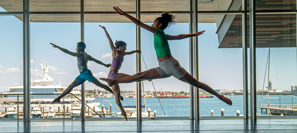Dancers leap in front of large windows facing the Boston harbor