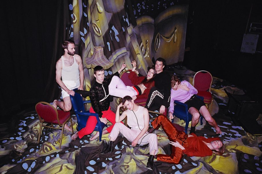 A group of people in colorful, eclectic clothing pose on a patterned floor with red chairs. The background is abstract with draped fabric and a lamp. The scene has a theatrical, artistic vibe.