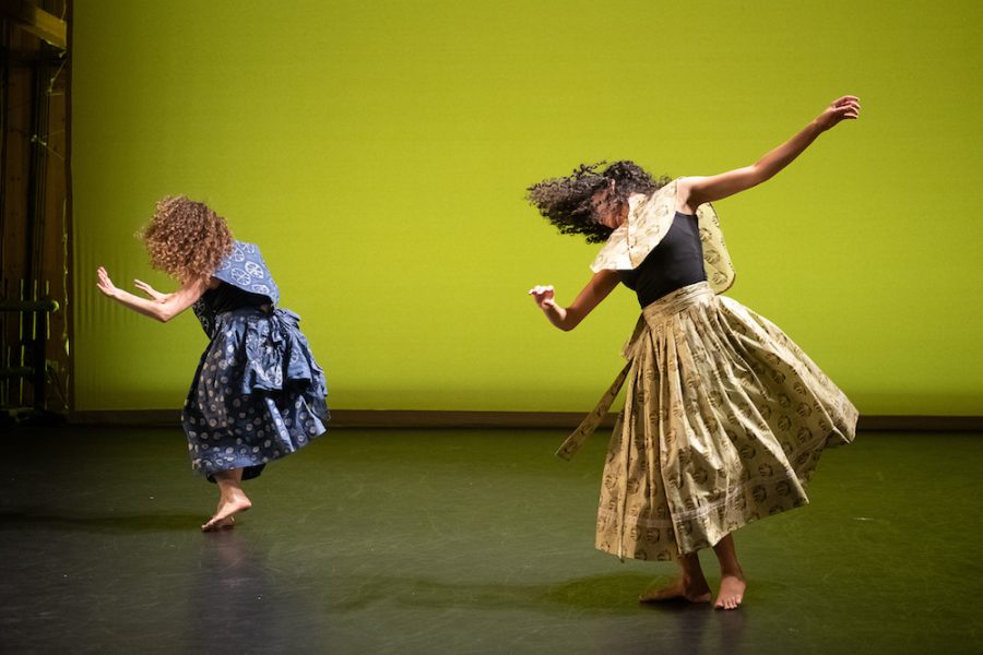 Two dancers with curly hair perform on stage, wearing skirts with intricate patterns. One skirt is blue, and the other is beige. They are captured mid-movement against a vibrant green background.