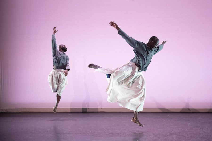 Two dancers perform mid-air leaps on stage, wearing flowing white skirts and blue tops. They appear energetic and poised against a soft pink backdrop, creating a sense of movement and grace.