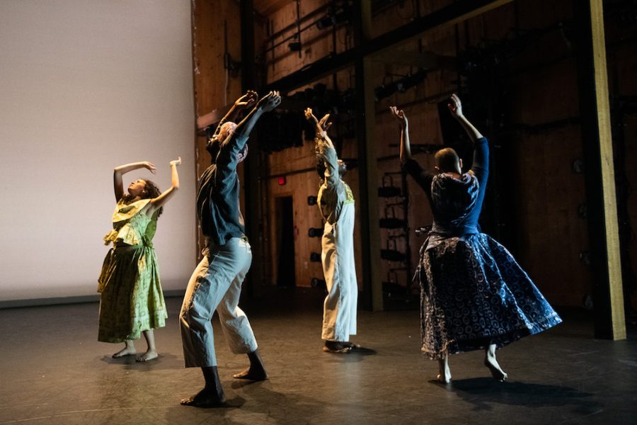 Four dancers perform on a dimly lit stage with a wooden backdrop. They are expressive, wearing casual and flowing costumes. Their dynamic poses and the lighting create a dramatic ambience.