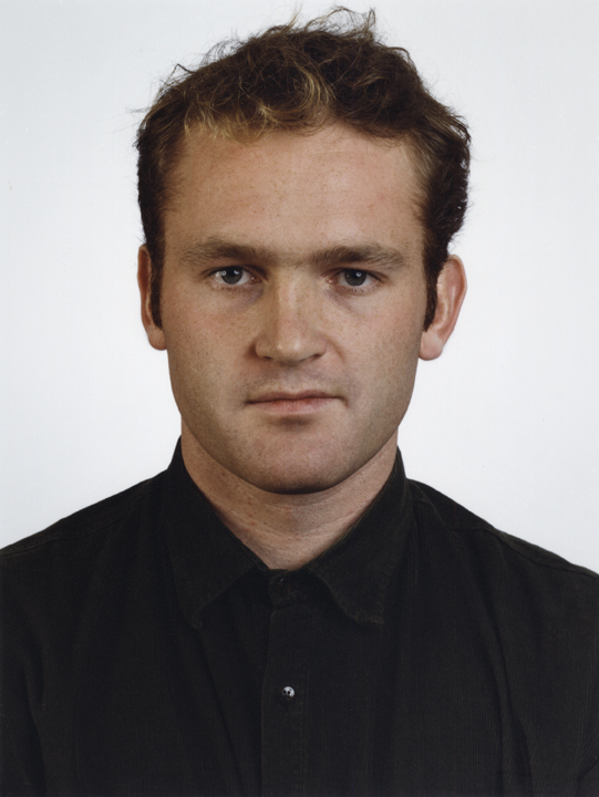 A color photograph portrait resembling an ID photo of a young man with light skin, dark hair, and a black button-up shirt, shown in close-up and staring directly at the viewer.