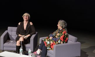 Two people are sitting on gray chairs on a stage. One person is laughing, wearing a black dress and knee-high boots. The other is wearing a floral-patterned coat and holding a microphone. A small table with water bottles is in front of them.