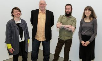 Four people stand side by side against a plain white background. Three are smiling, and one has a neutral expression. They are casually dressed, with varying styles, and each adopts a relaxed pose.