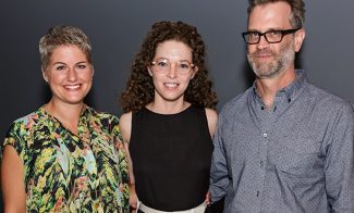 Three people stand together smiling against a dark background. The person on the left wears a colorful patterned top, the middle person wears glasses and a black dress, and the person on the right wears glasses and a blue button-up shirt.
