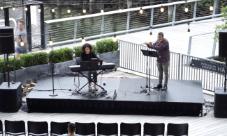 A small outdoor stage with two performers: one playing a keyboard and the other playing a flute. The stage is set on a wooden deck by water, surrounded by string lights and greenery. Rows of empty chairs face the stage.