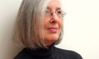A woman with shoulder-length gray hair and glasses, wearing a black turtleneck, looks thoughtfully to the side against a plain white background.
