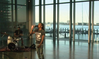 A singer performs energetically on stage with a microphone, accompanied by a drummer. The backdrop features large glass windows showcasing a marina and cityscape under a clear sky.