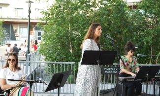 A woman singing into a microphone with a music stand in front of her, accompanied by two musicians playing keyboards and trumpet. They are performing outdoors on a stage surrounded by greenery and some onlookers in the background.