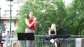 Two women performing outdoors: one sings into a microphone, wearing a red top and patterned pants; the other plays a keyboard, wearing a black dress. They are on a patio with trees in the background and people gathered nearby.