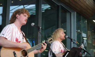 Two musicians with curly hair perform outdoors. The musician on the left plays an acoustic guitar and sings into a microphone, while the musician on the right also sings, with music stands in front. Both wear baseball jerseys with Phillies on them.