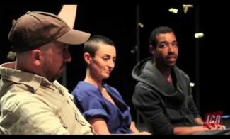 Three individuals are sitting in a dimly lit setting, engaging in a discussion. One wears a cap, another has a shaved head and smiles slightly, and the third person looks attentive. The letters ICA are visible in the bottom right corner.