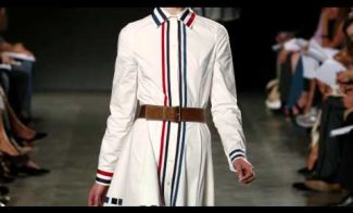 Model walking on a runway wearing a white shirt dress with vertical red and blue stripes and a brown belt. Audience members are seated along the sides of the runway.