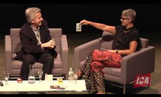 Two people sit on stage in armchairs during a discussion. The person on the right, wearing glasses and colorful pants, holds up a note, while the person on the left, wearing a suit, smiles. A table with papers and drinks is in front of them.