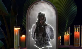 A glowing stone monument featuring a person with long braided hair, set against large leaves. Candles surround the monument, casting a warm light, creating an atmospheric scene.