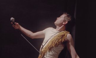 A person wearing a white shirt and a gold fringe across their shoulder holds a microphone, leaning back dramatically. The background is dimly lit, creating a spotlight effect on the person.