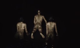 Three figures covered in gold paint pose dramatically against a dark background. The central figure is mid-air, while the others are crouched, creating a sense of motion and artistry. The lighting highlights their muscular forms.