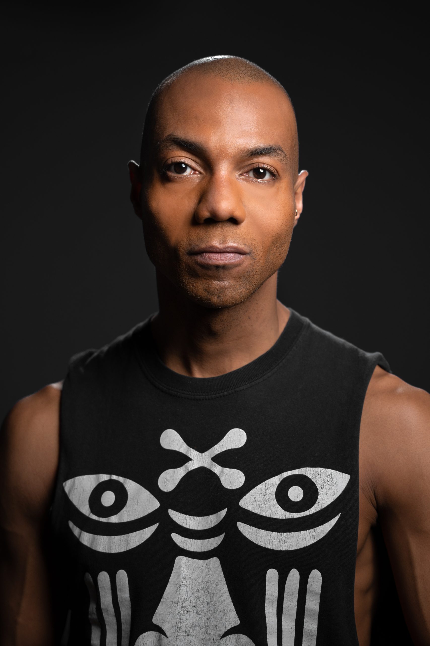 A person with a shaved head wearing a sleeveless shirt with a bold graphic design, featuring stylized eyes and shapes. The background is dark, and the person is looking directly at the camera with a calm expression.